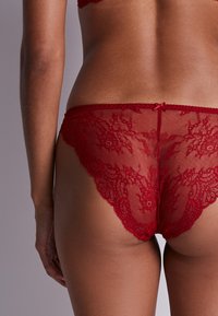 String en dentelle rouge avec des motifs floraux, des bords festonnés et un petit nœud à l'arrière ; texture translucide et délicate qui contraste avec le teint de la peau.