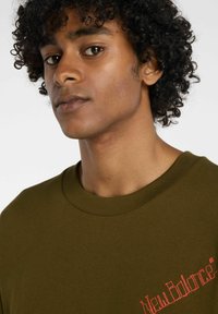 Olivegrünes Baumwoll-T-Shirt mit Rundhalsausschnitt, mit rot gesticktem "New Balance"-Text auf der linken Seite. Glatte Textur.