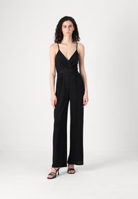 Crna kombinezon s dubokim V-izrezom, omotanim prednjim dijelom i širokim nogavicama. Ima tanke naramenice i mekanu teksturu, uparen s crnim sandalama sa trakama.