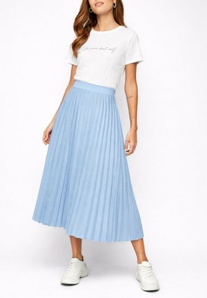 Femme portant une chemise blanche à manches courtes avec texte, jupe-midi plissée bleu clair, baskets blanches, debout devant un fond blanc.