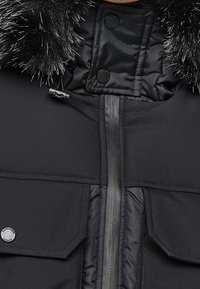 Manteau d'hiver noir avec fermeture éclair centrale, deux poches à rabat avec boutons-pression, et capuche bordée de fourrure avec cordon de serrage.
