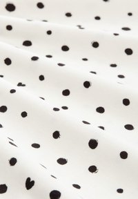 Schwarze Polka-Dots auf einem weißen Stoff erzeugen ein texturiertes Muster mit variierenden Punktgrößen und einer glatten, weichen Erscheinung.