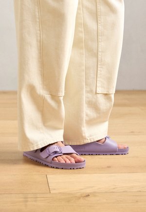 Stopy w jasnofioletowych sandałach na otwarty palec z zapięciem, stojące na drewnianej podłodze, w zestawieniu z szerokimi, kremowymi spodniami.