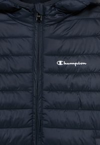 Μπουφάν puffer σε ναυτικό μπλε, με φερμουάρ, ραμμένες ενότητες και λευκό λογότυπο Champion στην αριστερή πλευρά. Ομαλή, γυαλιστερή υφή.