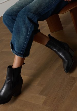 Zwarte leren enkellaarsjes met een spitse neus en een blokhak, gedragen met opgerolde blauwe spijkerbroeken op een houten vloer.