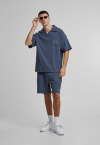 Mørkeblå kortermet skjorte med lomme og brodert logo, iført med matchende shorts. Hvite joggesko. Glatt stofftekstur.