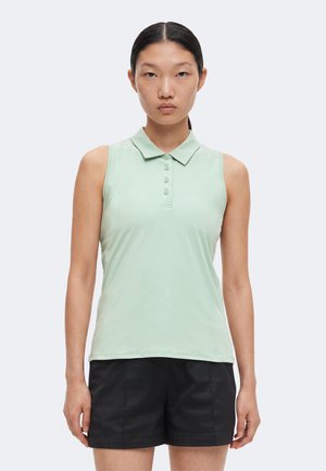 Femme aux cheveux noirs et raides portant un polo vert clair sans manches et un short noir, debout face à l’avant sur un fond uni.