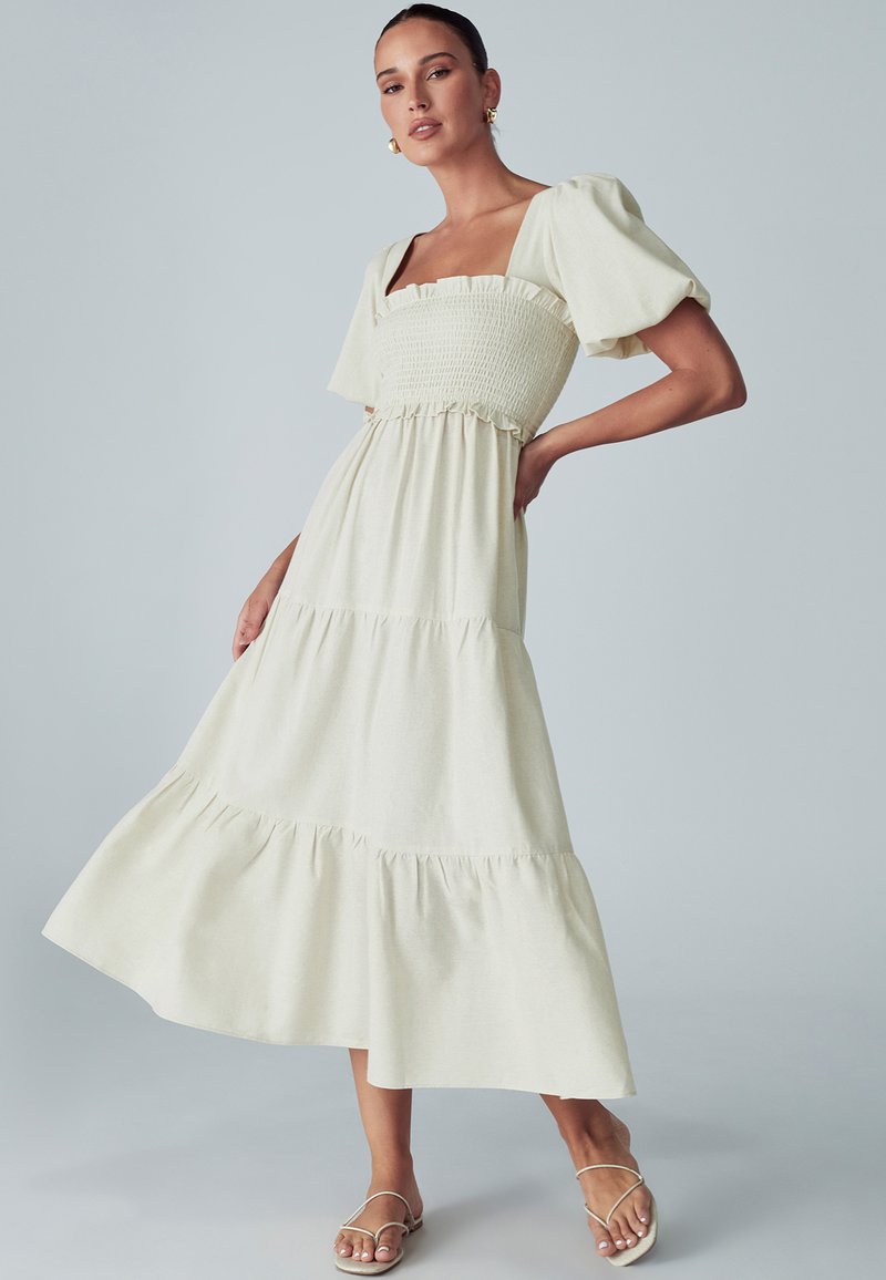 Šviesiai bežinis midi suknelė su trumpomis pūstomis rankovėmis, kvadratiniu iškirpimu, surinkta krūtine ir sluoksniuotais volanais. Minkšta, sklandi audinio tekstūra.