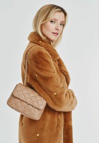 Beżowa torebka pikowana z złotym łańcuszkiem, trzymana przy brązowym płaszczu z sztucznego futra z złotymi guzikami. Prosty i elegancki design.