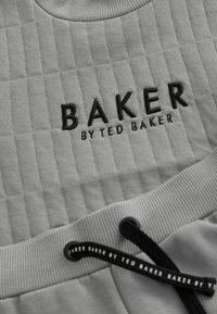 Šedá atletická tkanina s prešívanou textúrou. Logo "BAKER BY TED BAKER" čierne vyšívanie. Obsahuje šnúrkový uzáver a rebrované akcenty.