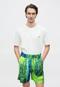 Weißes Baumwoll-T-Shirt kombiniert mit lebhaft grün-blau gemusterten Badeshorts mit elastischem Bund und Seitentaschen.