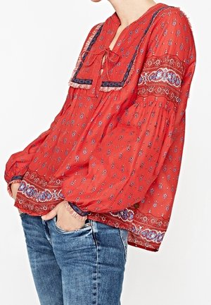 Losse rode blouse met blauw-witte bloemmotieven, decoratieve vierkante halslijn met franjes, gecombineerd met een blauwe spijkerbroek.