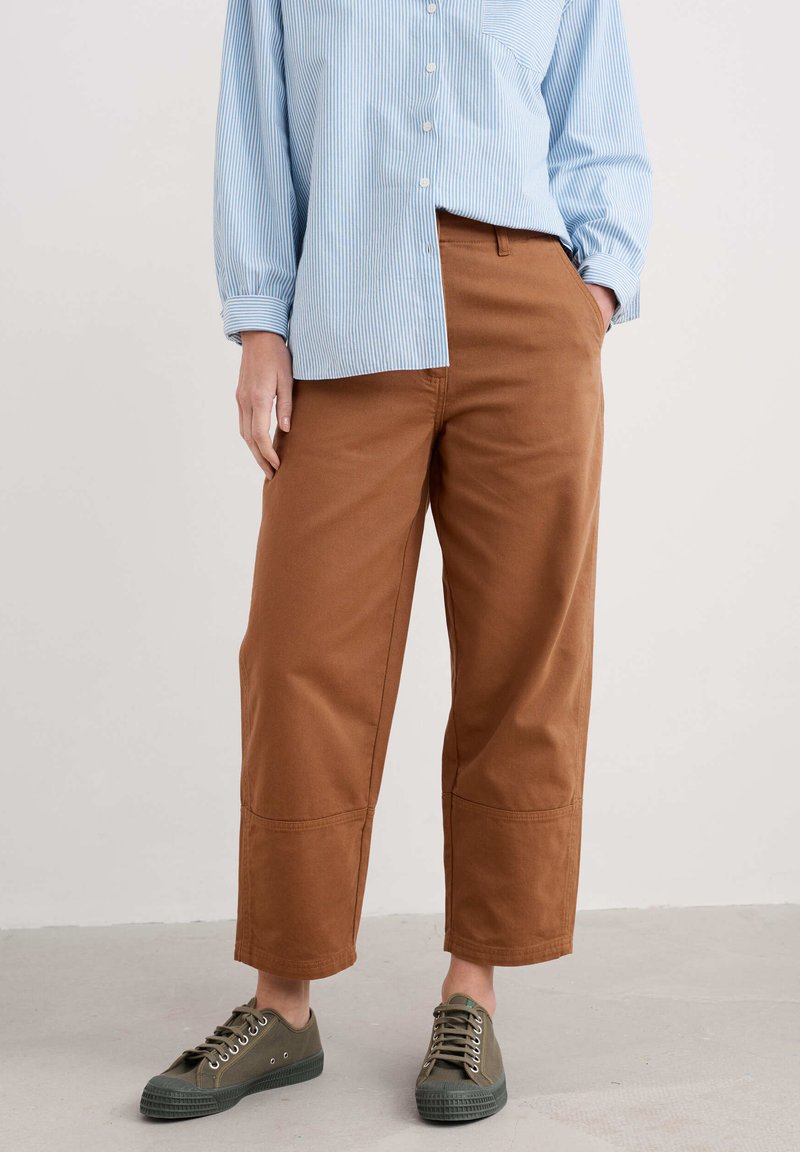 Bruine, wijde broek met een gladde textuur, voorzien van zijzakken en een gestikte zoom, gedragen met olijfgroene canvas sneakers.