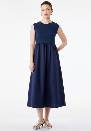 Kvinde står iført en ærmeløs midi-kjole i navyblå med en indsnøret talje og hvide åbne hælede sandaler på hvid baggrund.