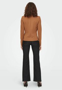 Veste marron sur mesure avec épaules structurées et coupe ajustée, associée à un jean évasé foncé et des chaussures noires à talons hauts.