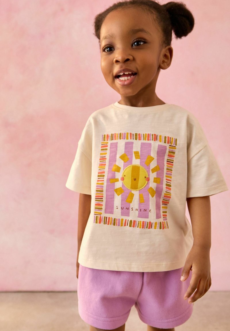 Jeune fille avec deux couettes bouffantes portant un t-shirt crème avec un soleil coloré et le texte "Sunshine", et un short rose sur un fond rose.