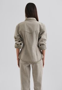 Harmaa denimbomber, jossa on terävät kaulukset, käännetyt hihat ja slim fit -malli. Takana on yoke-sauma ja ompeluyksityiskohtia.