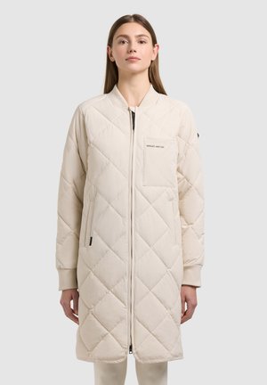 Femeie purtând o geacă lungă matlasată bej, cu fermoar frontal, buzunar la piept și buzunare laterale, în picioare pe un fundal simplu.