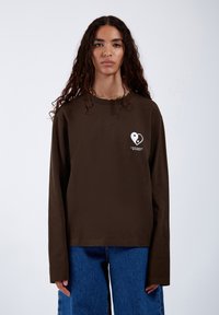 Braunes Langarmshirt aus Baumwolle mit rundem Halsausschnitt, auf der linken Brust mit einem weißen Herzlogo und Text. Kombiniert mit blauen Jeans.