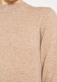 Pull en maille marron clair avec une texture douce. Il présente un col rond et des manches montées, mettant en valeur un motif chiné subtil.
