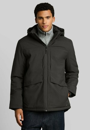 Young man wearing a black hooded winter jacket over a gray zip-up sweater, standing with one hand in jacket pocket against a gray background.