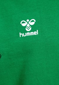 T-shirt in cotone verde con logo bianco "hummel" e design di un'ape. Tessuto morbido con cuciture visibili e maniche corte.