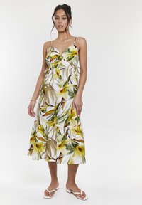 Vestido midi floral con tirantes finos, que presenta un patrón de hojas verdes y amarillas sobre un fondo blanco; bajo con volantes y lazos laterales.