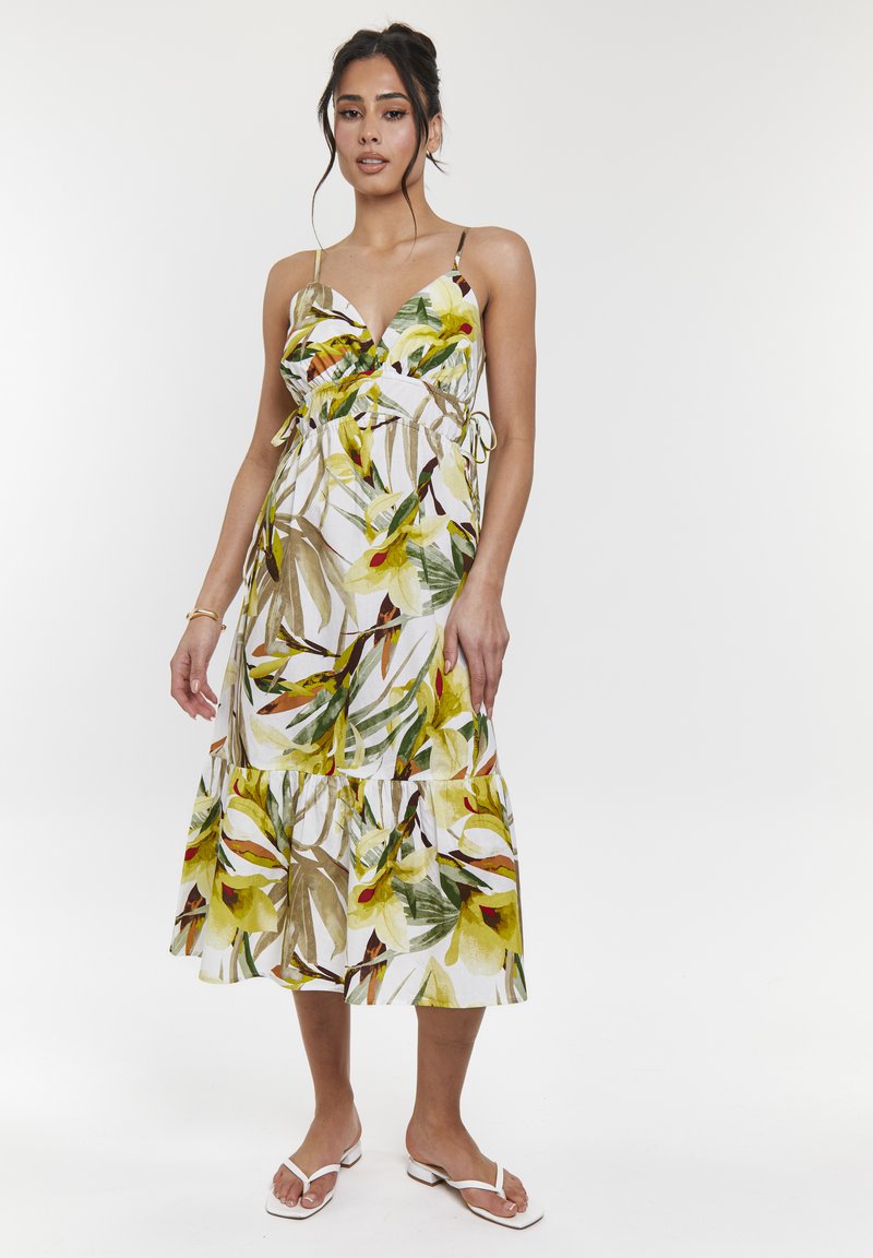 Vestido midi floral con tirantes finos, que presenta un patrón de hojas verdes y amarillas sobre un fondo blanco; bajo con volantes y lazos laterales.