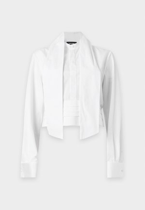 Femme portant un chemisier blanc à manches longues avec un panneau avant et un pantalon noir à jambes larges, debout avec une main dans la poche sur fond blanc.