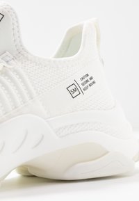 Valkoinen urheilujalkine, jossa on teksturoitu neulottu päällinen, pehmustettu nilkkareuna ja sileä kumipohja. Sivussa on varoitustarra.