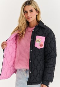 Žena v světle modrých džínách a růžovém chlupatém svetru ukazuje vnitřek černé prošívané bundy s růžovou podšívkou a kapsami.