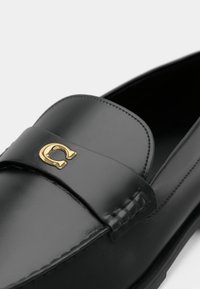 Mocassin en cuir noir avec bout arrondi, détails cousus et petit emblème métallique doré en forme de "C" sur la bride traversant le dessus.