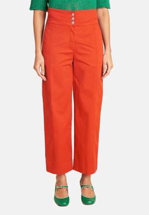 Frau trägt hoch taillierte, weit geschnittene orange Hose mit drei vorderen Knöpfen, grünes gemustertes Oberteil und grüne Mary Jane Schuhe.