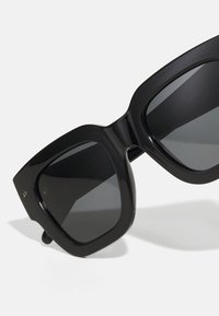 Storebriller i svart med bred ramme, flate linser og vinklet form. Den bemerkelsesverdige designen inkluderer glatte detaljer og minimal merkevare.
