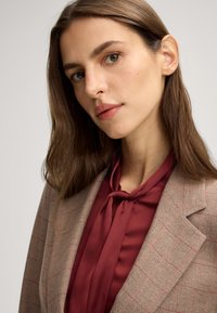 Blazer beige a motivi houndstooth con accenti rosa, abbinato a una blusa in satin bordeaux con cravattino al collo. Tessuto morbido, vestibilità sartoriale.