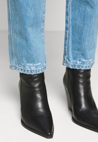 Jean bleu clair étiqueté "Pedal Pusher" au-dessus de bottines noires à bout pointu avec talon compensé sur une surface claire.