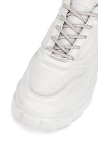 Zapatilla blanca con parte superior texturizada con perforaciones, cordones planos de color gris claro y suela blanca gruesa con elementos de diseño en capas.