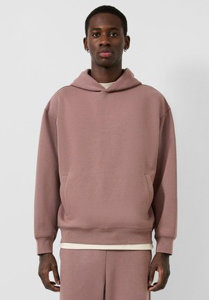 Jonge man die een mauve hoodie en bijpassende broek draagt, staand tegen een effen lichte achtergrond, naar voren kijkend.