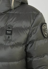 Graue Steppjacke mit glänzender Oberfläche, ausgestattet mit einem Logo-Patch und Druckknopfverschlüssen. Das Design umfasst genähte Paneele für einen strukturierten Look.