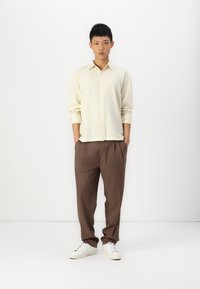 Light yellow button-up shirt, brown pleated pants, and white sneakers. The shirt has long sleeves and a relaxed fit. Plain colors, no patterns.
