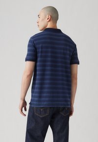 Cămașă polo bleu marin cu dungi, cu mâneci scurte și guler clasic, asortată cu blugi închis la culoare. Are un tiv drept și slits laterale.