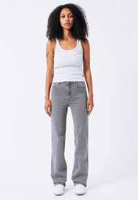 Grijze geribbelde tanktop gecombineerd met hoge grijze wijde jeans. Model staat in witte sneakers voor een effen achtergrond.