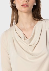 Beige Bluse mit Drapierung und Schalkragen. Glatte, weiche Textur. Das Model trägt geometrische silberne Ohrringe. Fokus auf dem Oberkörper und dem Halsauschnitt.