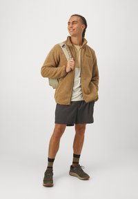 Brun fleecejakke, tan baselag, grå shorts. Merknader: teksturert stoff, lett ryggsekk, og mørkt fottøy med kontrasterende sokker.