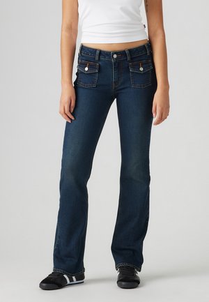 Femme portant un jean bootcut bleu foncé avec des poches à rabat à l'avant et un débardeur court blanc, debout devant un fond uni.