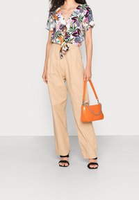 Blomstret kortærmet bluse med bindefront, kombineret med højtaljede beige bukser. Accessoriseret med en orange håndtaske og sorte sandaletter med hæl.