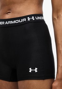 Κοντινό πλάνο ενός ατόμου που φοράει μαύρα σορτς συμπίεσης Under Armour, εμφανίζοντας το λογότυπο στη μέση και στην περιοχή του μηρού.