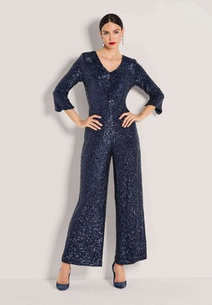Femme portant une combinaison bleu marine à manches longues pailletée avec jambes larges, associée à des talons bleu marine et des boucles d'oreilles remarquables, posant les mains sur les hanches.