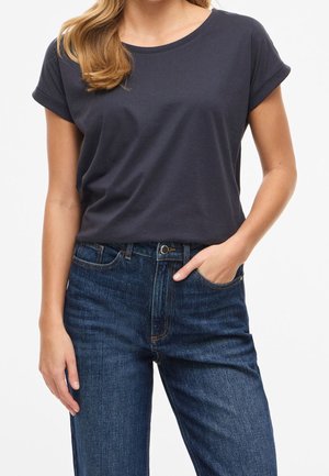 Vrouw die een marineblauw T-shirt met korte mouwen draagt, ingestopt in een hooggetailleerde donkerblauwe jeans met één hand in de zak.