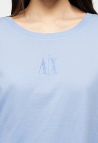 Γυναίκα που φοράει γαλάζιο μπλουζάκι με στρογγυλή λαιμόκοψη και μικρό λογότυπο "A|X" από στρας στο κέντρο του στήθους.